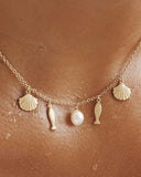 Gold plated charm necklace with shell, fish and natural pearl pendants on a delicate chain, inspired by Mediterranean summer style — επιχρυσωμένο κολιέ με charms κοχύλι, ψαράκι και φυσικό μαργαριτάρι σε λεπτή αλυσίδα, εμπνευσμένο από το καλοκαιρινό μεσογειακό στυλ. Διαθέσιμο στο Iconic Angels Glyfada.