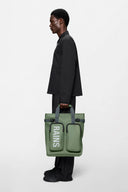 Waterproof green backpack with bold RAINS logo, structured design and practical front pockets for everyday essentials — αδιάβροχο πράσινο backpack με statement λογότυπο RAINS, minimal αισθητική και λειτουργικές μπροστινές θήκες για άνετη οργάνωση στην πόλη ή στις αποδράσεις του weekend. Διαθέσιμο στο Iconic Angels Glyfada.