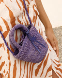Mini raffia summer bag in purple with braided texture and top handle, styled crossbody over printed linen dress — μικρή καλοκαιρινή τσάντα από ψάθα σε μωβ απόχρωση με πλεκτή υφή και χερούλι, ιδανική για city & resort looks. Διαθέσιμη στο Iconic Angels Glyfada.